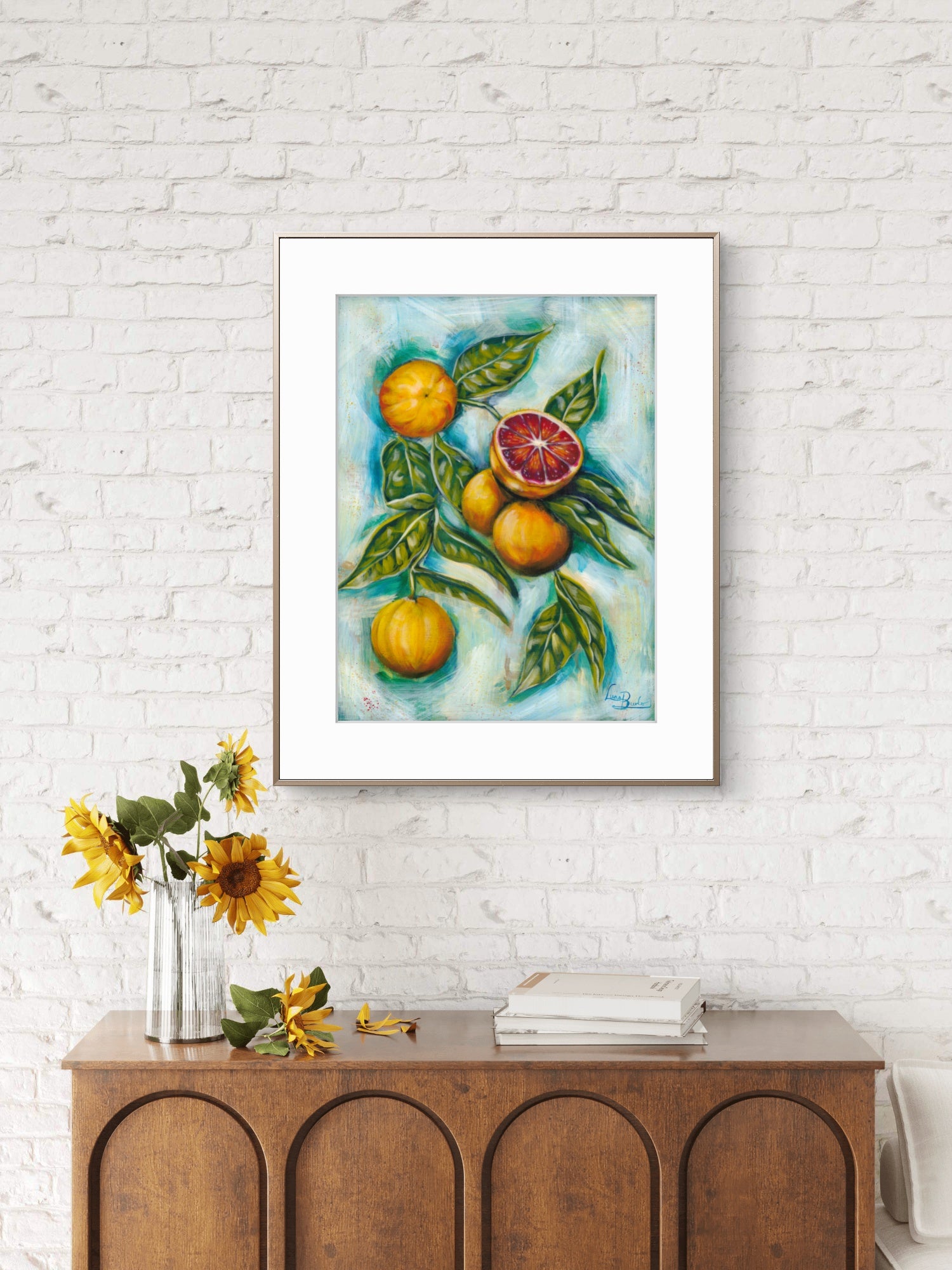 Framed Luna Burlo artwork of fruits and leaves on a white brick wall above a wooden console table with decorative items.
