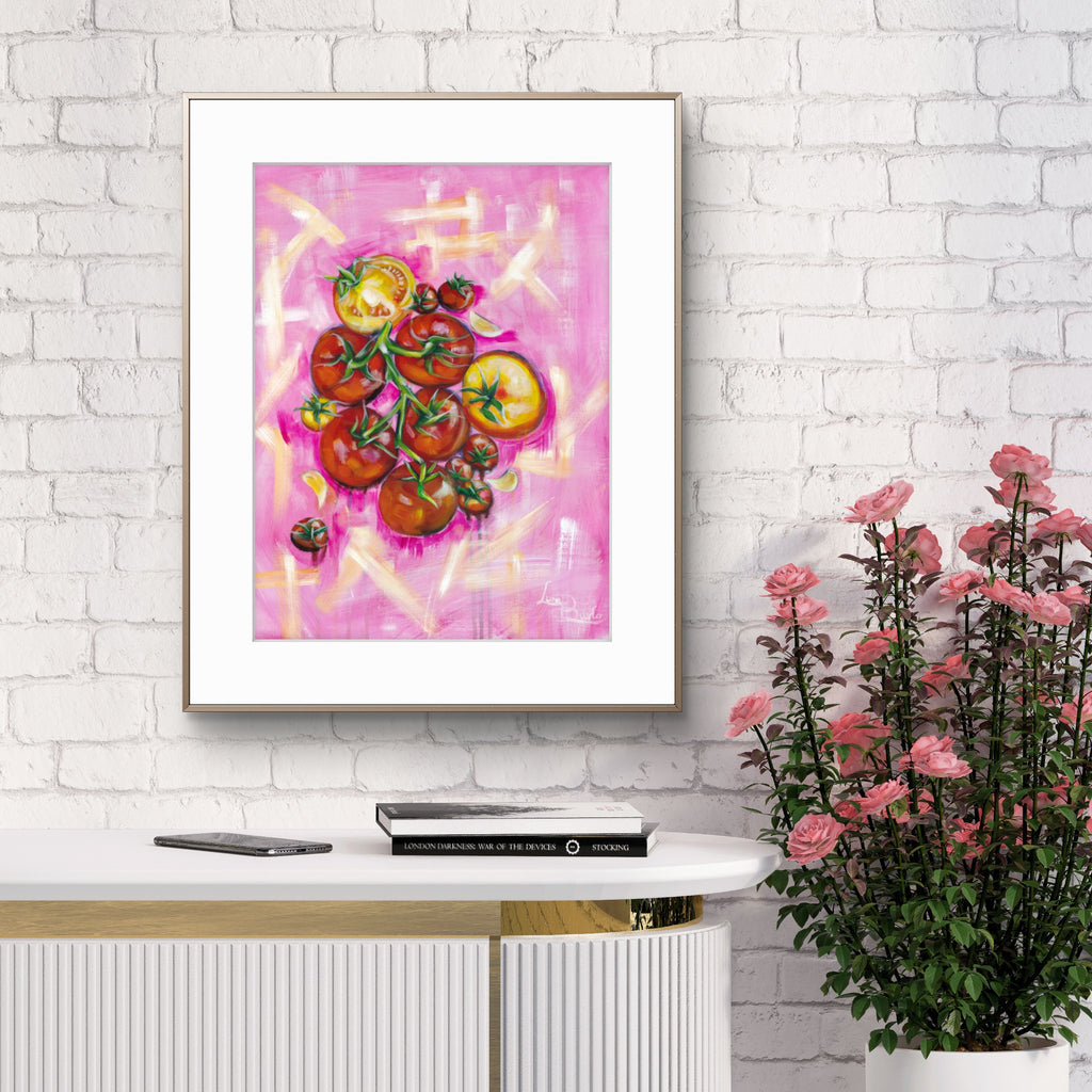 Framed Luna Burlo artwork of tomatoes on a pink background, displayed on a white brick wall above a white cabinet with a plant to the right.