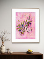 Framed Luna Burlo artwork with olives design on a pink background, displayed on a wall above a wooden shelf.