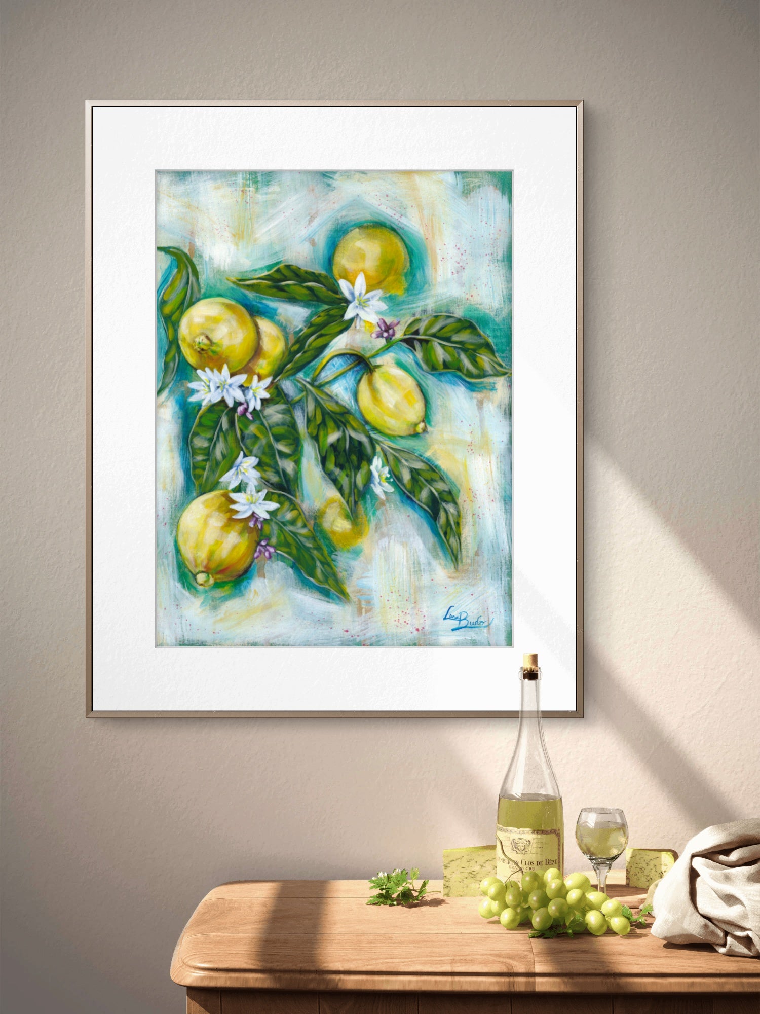 Framed painting of lemons and leaves on a wall above a table with wine and grapes. By Luna Burlo