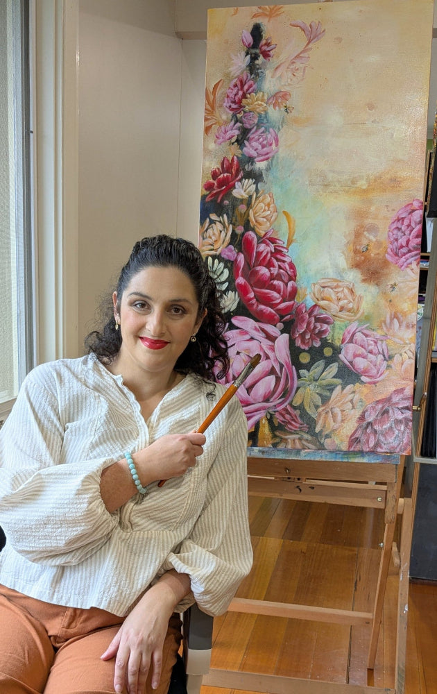 Woman holding a paintbrush in front of a colorful floral painting on an easel.