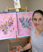 Luna Burlo holding a paintbrush in front of two Mediterranean Table Series paintings on an easel.