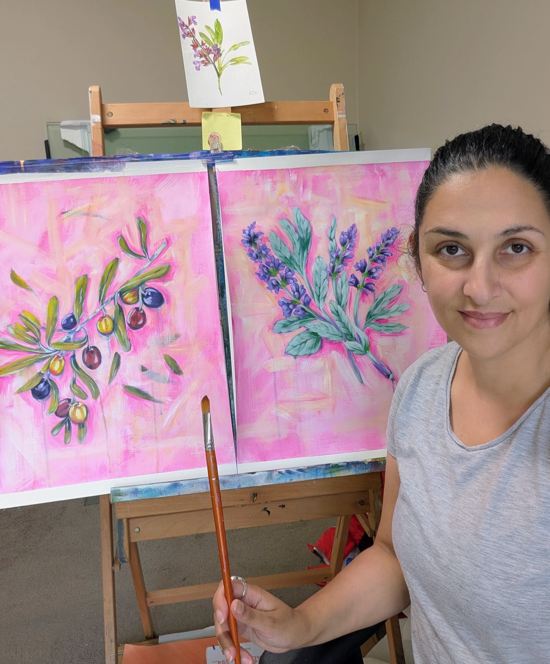 Luna Burlo holding a paintbrush in front of two Mediterranean Table Series paintings on an easel.