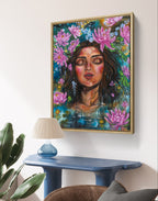 Framed abstract artwork of a woman's face surrounded by pink water lily flowers on a blue background in a room with a lamp and plant. Artwork by Luna Burlo, Melbourne, Australia