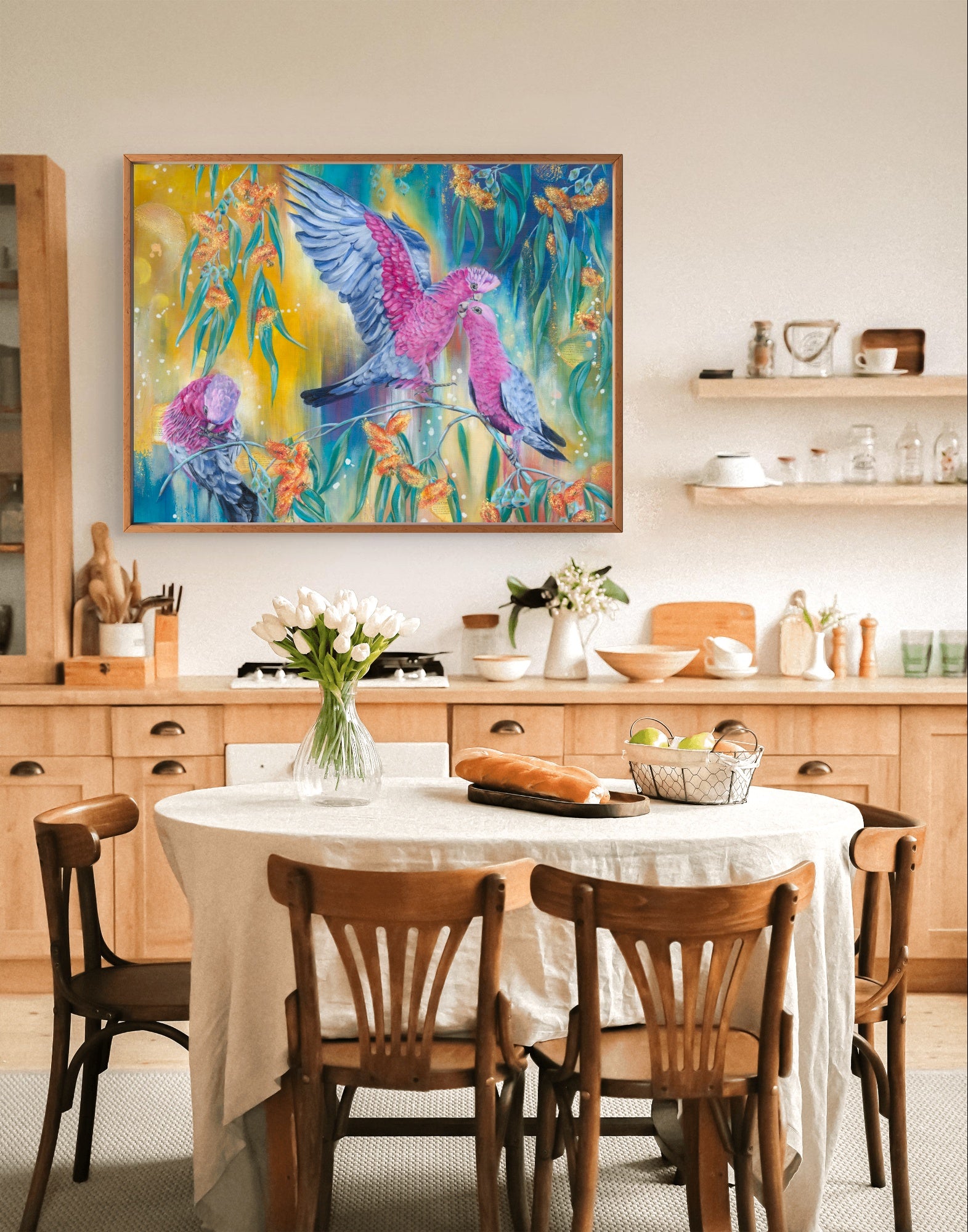 Dining room and kitchen with a colorful abstract fine art canvas print of a painting of native Australian galah birds and native flowers on the wall above a table and white walls. Painting is large, rectangle and in horizontal landscape orientation. Art by Luna Burlo, Melbourne, Australia