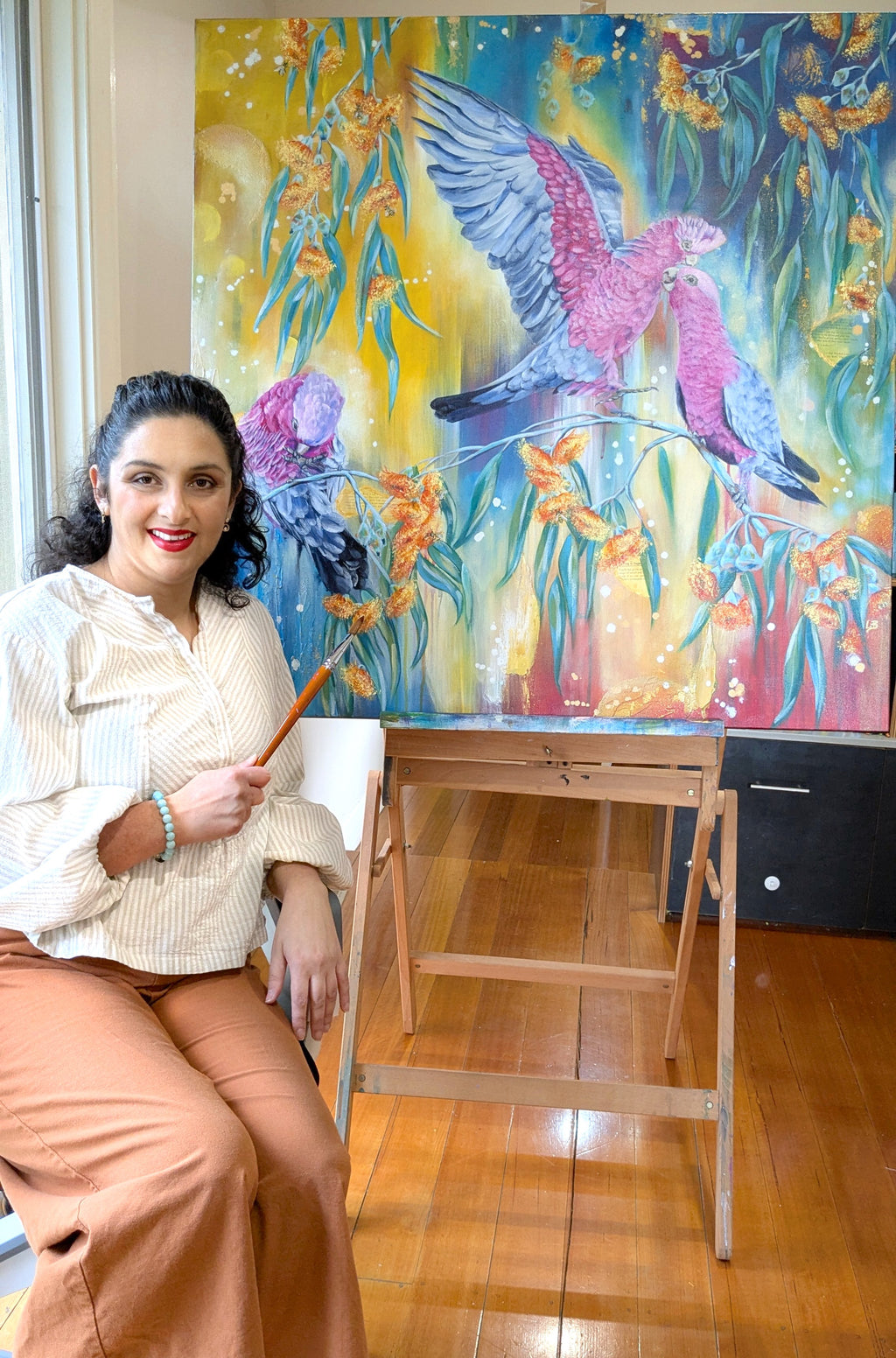 Melboure, Australia artist, Luna Burlo, sitting next to her colorful mixed media painting of pink native galah birds and eucalyptus tree blossom flowers on an easel.