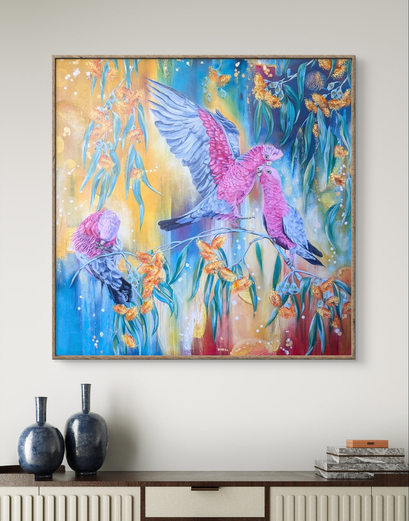 Colorful painting or fine art print of pink galahs, Native Australian parrots or birds on gum trees branches with flower blossoms in a wooden frame, displayed on a wall above a console table. Artist, Luna Burlo, Melbourne, Australia
