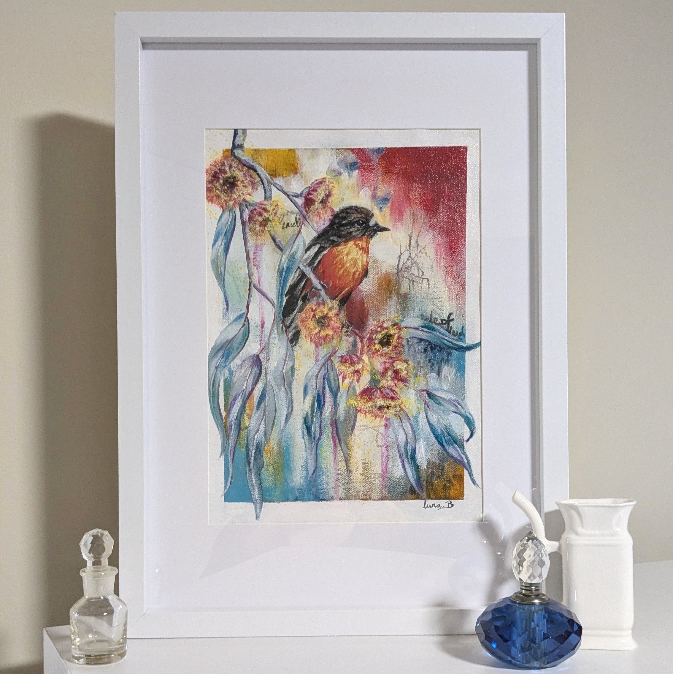 Framed Luna Burlo artwork of a Flame Robin bird and flowers on a white surface with decorative items.