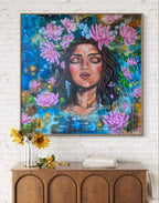 Colorful painting or fine art print of a woman's face surrounded by lotus and lukspur flowers on a white brick wall above a wooden console table. Birth flowers and celestial art for brith month of July, Leo, and Cancer. By Luna Burlo, Australia 