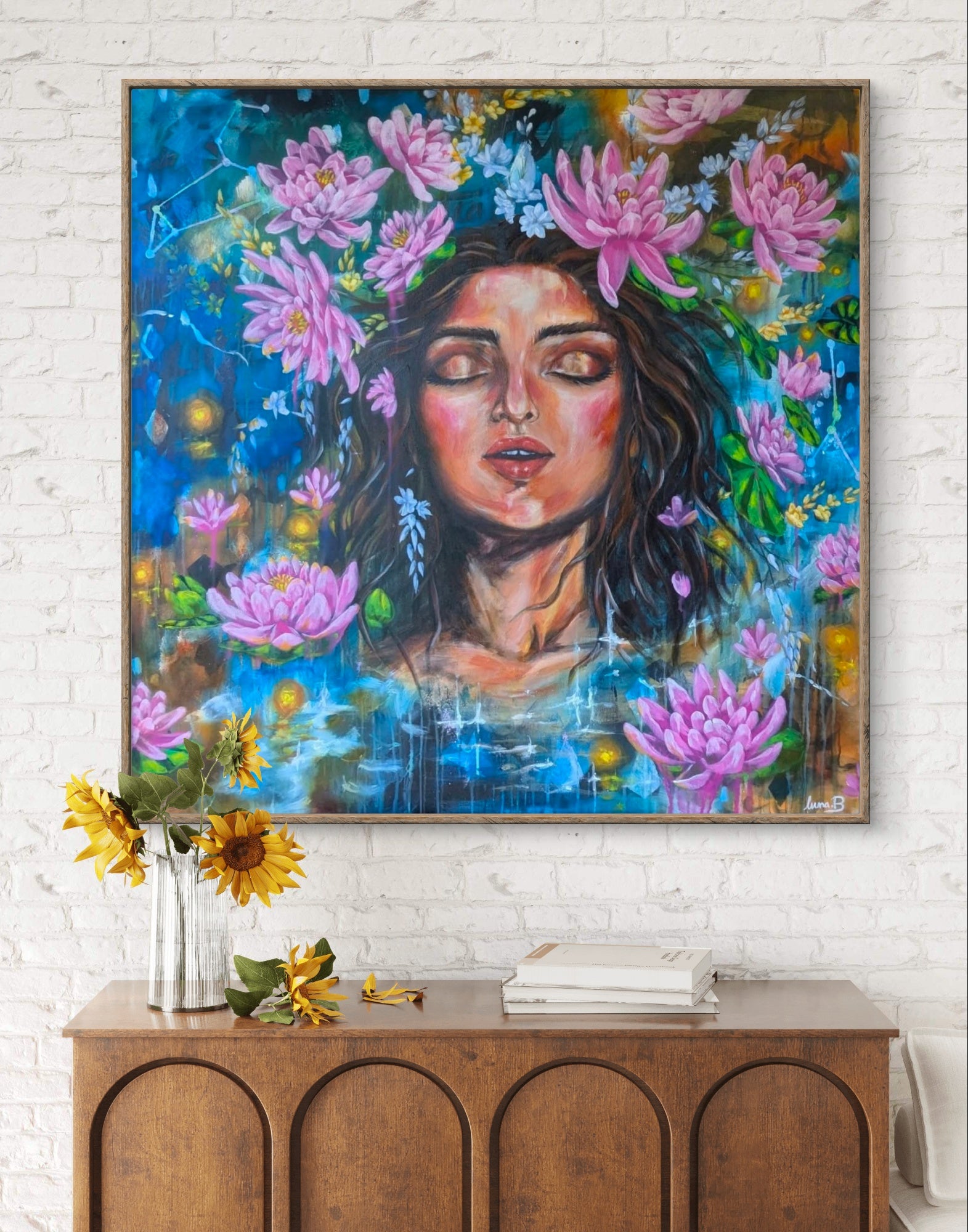 Colorful painting or fine art print of a woman's face surrounded by lotus and lukspur flowers on a white brick wall above a wooden console table. Birth flowers and celestial art for brith month of July, Leo, and Cancer. By Luna Burlo, Australia 