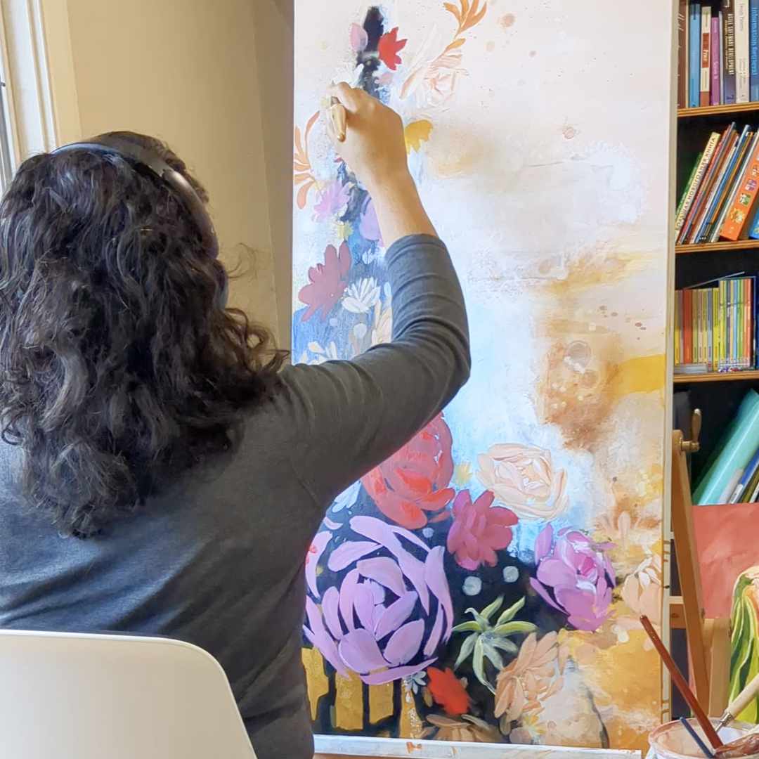 Luna Burlo painting a colorful floral scene on a large canvas with books and art supplies in the background.