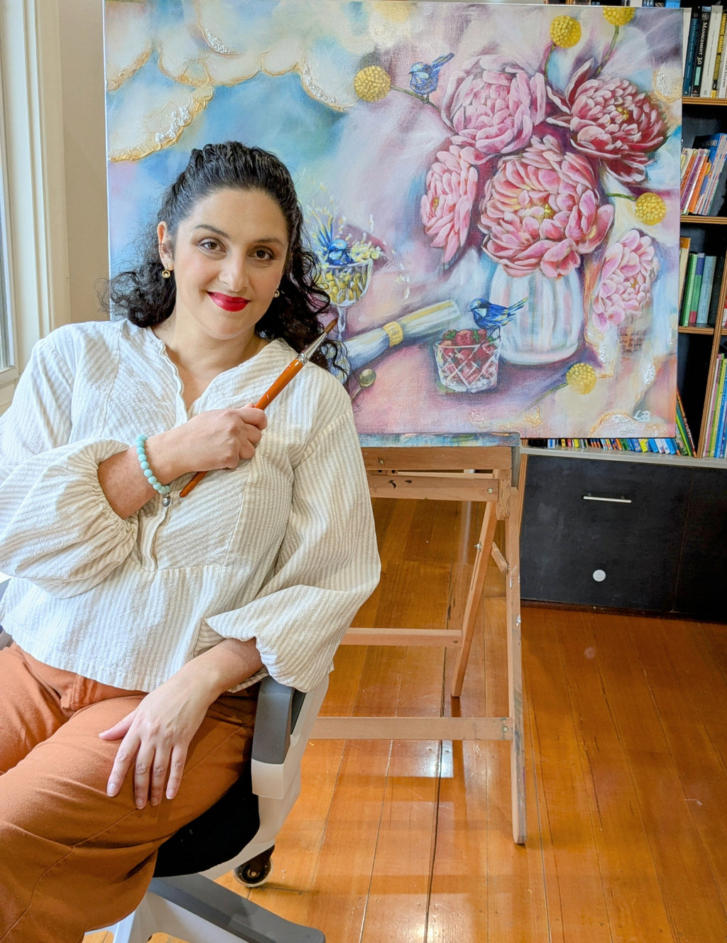 Artist, Luna Burlo, holding a paintbrush in front of her painting of pink peony flowers in in her Melbourne, Australia art studio.